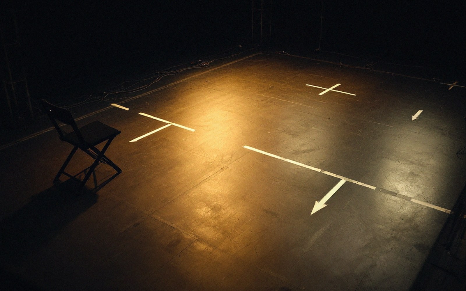 Empty stage floor with taped marks and arrows lit by a single spotlight beside a folding chair